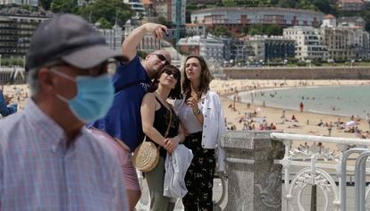 Un señor con mascarilla pasa por delante de un grupo de turistas que se fotografían en la playa donostiarra de La Concha.