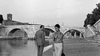 A man and a woman exchange timid glances by a river in Italy circa the 1960s.
