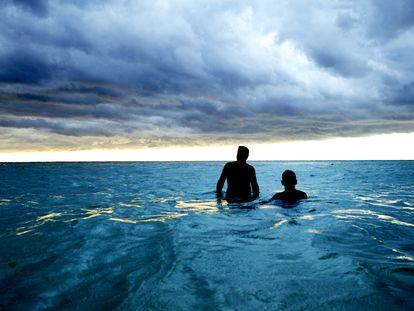 Two men take a dip in the ocean in Cuba, with storm clouds approaching.