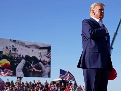 As footage from the Jan. 6, 2021, insurrection at the U.S. Capitol is displayed in the background, former President Donald Trump stands while a song, "Justice for All," is played during a campaign rally at Waco Regional Airport, Saturday, March 25, 2023, in Waco, Texas.