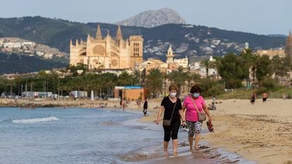 Two women wear masks walk as they take a walk on a beach in Mallorca.