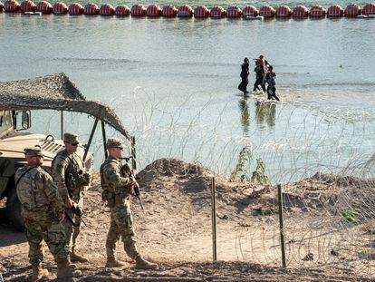 Asylum-seeking migrants walk in the Rio Grande river between a floating fence and the river bank as they look for an opening in a concertina wire fence to land on U.S. soil in Eagle Pass, Texas